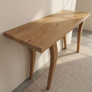 Op de afbeelding: Een lichtbruine houten consoletafel met een rechthoekig blad en gebogen poten. De tafel is gemaakt van hout en heeft een natuurlijke afwerking. De tafel is ongeveer 122 cm lang, 30 cm breed en 76 cm hoog.