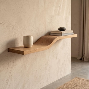 May include: A stylish, floating wooden shelf with a curved design. The shelf is made of light-coloured wood and is mounted on a beige wall. A ceramic cup, two books, and a dark stone are displayed on the shelf.