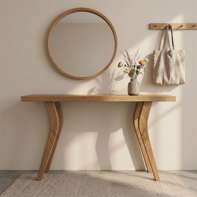 Op de afbeelding: Een houten console met gebogen poten, een ronde spiegel en een beige draagtas aan de muur. Een kleine vaas met bloemen staat op de tafel. De tafel is gemaakt van lichtgekleurd hout.