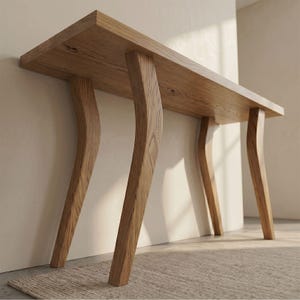 Op de afbeelding: Een lichtbruine houten console met een rechthoekig blad en vier gebogen poten. De tafel is gemaakt van massief hout met een natuurlijke afwerking. De poten zijn schuin geplaatst en zorgen voor een uniek design. De tafel staat tegen een neutrale muur.