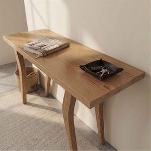Op de afbeelding: Een lichtbruine houten console met een rechthoekig blad en gebogen poten. Een boek en een klein leren dienblad met sleutels liggen op de tafel. Een gevlochten mand is zichtbaar op de achtergrond.