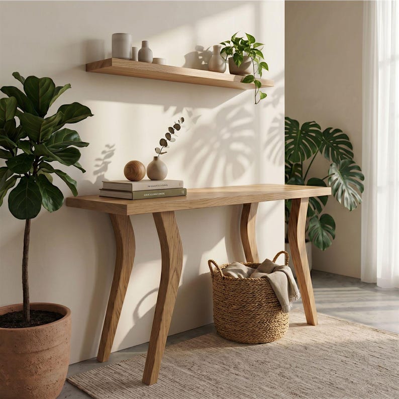 Op de afbeelding: Een lichtgekleurde houten console met gebogen poten, een plank en een gevlochten mand. De tafel is versierd met boeken, vazen en decoratieve items. Een potplant staat links en een vloerkleed ligt op de vloer.