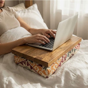 May include: A wooden laptop desk with a patterned fabric base. The desk has a light brown wood finish. The fabric base features a geometric design in orange, red, and blue. A person is using a laptop on the desk while lying in bed.