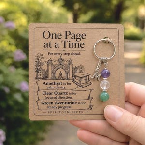 May include: A silver-toned keychain featuring a letter M charm and three beads: a purple amethyst, a clear quartz, and a green aventurine. The keychain is attached to a rectangular tan card printed with the text One Page at a Time and descriptions of the stones.