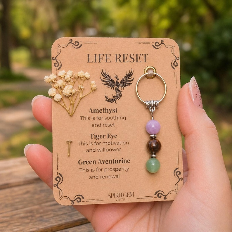 May include: A keychain with three gemstone beads in purple, brown, and green, attached to a silver ring. The keychain is displayed on a brown card with the text "LIFE RESET" and the names of the gemstones. The card also features a phoenix illustration and dried flowers.