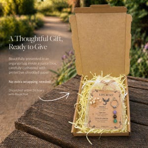 May include: An open cardboard gift box on a wooden surface, containing a Life Reset keychain with amethyst, tiger eye, and green aventurine beads. The box includes shredded paper, a card, and a small organza bag.