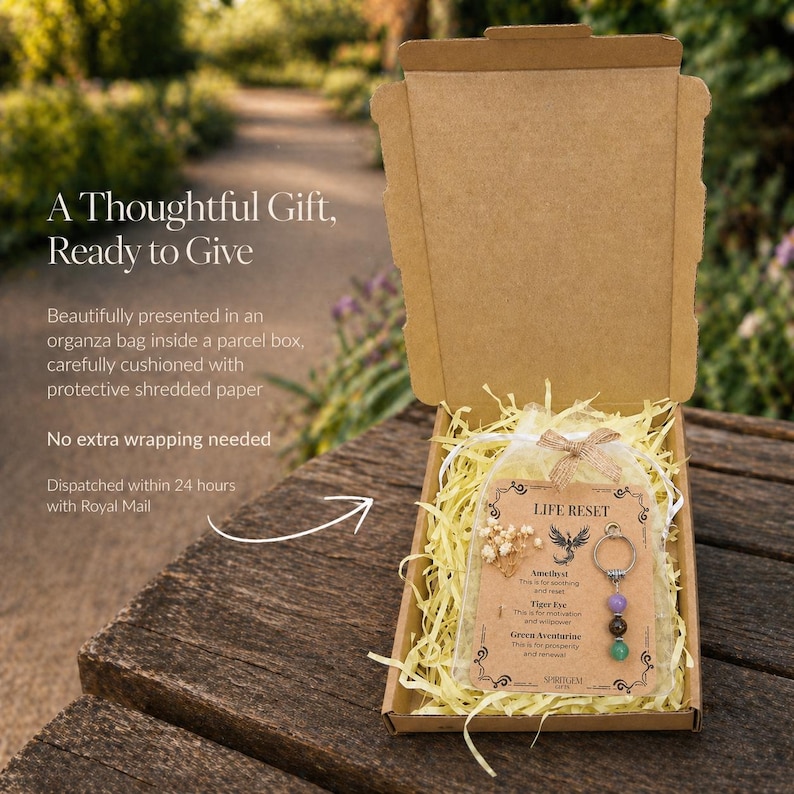 May include: An open cardboard gift box on a wooden surface, containing a Life Reset keychain with amethyst, tiger eye, and green aventurine beads. The box includes shredded paper, a card, and a small organza bag.