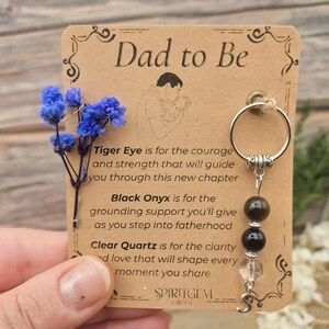 May include: A keychain with a brown card that reads "Dad to Be" and a drawing of a father holding a baby. The keychain has a silver ring with three beads: tiger eye, black onyx, and clear quartz. Blue flowers are attached to the card.