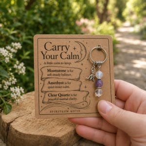 May include: A silver-toned metal keyring attached to a rectangular kraft paper card. The card features the text Carry Your Calm and descriptions of moonstone, amethyst, and clear quartz. The keyring includes a letter M charm and three beads.