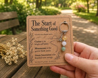 May include: A silver-toned keychain featuring a letter M charm and three round gemstone beads in yellow, white, and green, attached to a rectangular kraft paper card printed with the text The Start of Something Good and descriptions of citrine, moonstone, and green aventurine.
