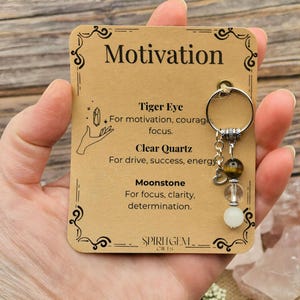 Puede incluir: Un llavero con un anillo plateado y tres cuentas de piedras preciosas: ojo de tigre, cuarzo transparente y piedra lunar. El llavero cuelga de una tarjeta marrón con la palabra "Motivation".