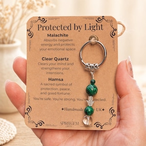 Könnte beinhalten: Ein Schlüsselanhänger mit einem silbernen Ring und einem Anhänger mit grünen Malachitperlen, einer klaren Quarzperle und einer silbernen Hamsa-Hand. Die Karte trägt die Aufschrift "Protected by Light" und listet die Eigenschaften der Steine auf.