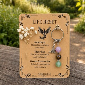 May include: A keychain with a silver ring and three gemstone beads: amethyst, tiger eye, and green aventurine. The keychain hangs from a brown card with the text "LIFE RESET" and "SPIRITGEM GIFTS". The card also features a phoenix illustration.