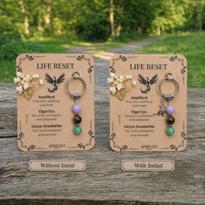May include: Two keychains with gemstone beads, amethyst, tiger eye, and green aventurine, are displayed on decorative cards. The cards read "LIFE RESET" and include text about the gemstones' properties. One keychain has an initial charm.