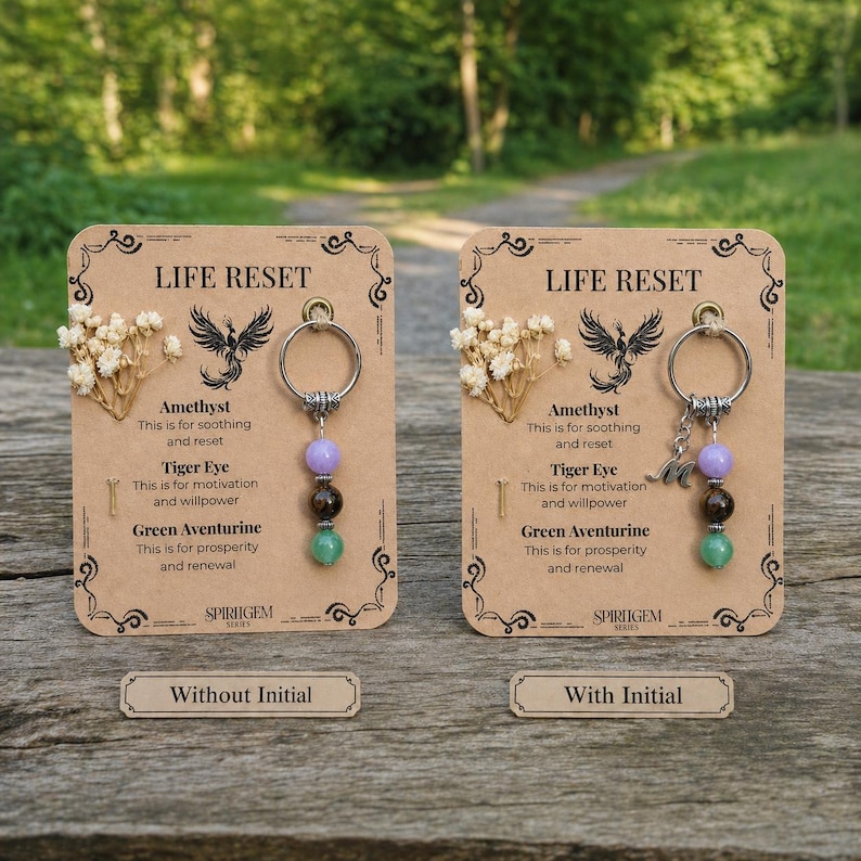 May include: Two keychains with gemstone beads, amethyst, tiger eye, and green aventurine, are displayed on decorative cards. The cards read "LIFE RESET" and include text about the gemstones' properties. One keychain has an initial charm.