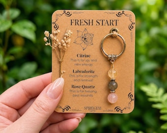 May include: A gold-toned metal ring keychain featuring three hanging gemstone beads: a pale yellow citrine, a grey labradorite, and a pink rose quartz. The keychain is attached to a rectangular tan card labeled FRESH START.