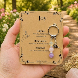 Puede incluir: Un llavero con un anillo plateado y tres cuentas de piedras preciosas: citrino amarillo, cuarzo rosa y amatista púrpura. El llavero está unido a una tarjeta que dice "Joy" y enumera las propiedades de cada piedra.