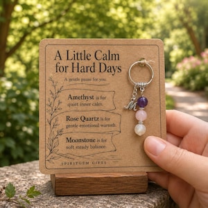 May include: A silver-toned metal keyring featuring a dangling charm with a letter M and three round beads in purple, light pink, and white, attached to a rectangular cardboard display card with the text A Little Calm for Hard Days.