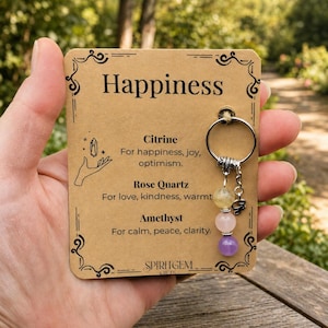 Puede incluir: Un llavero con tres cuentas de piedras preciosas: citrino amarillo, cuarzo rosa y amatista morada. El llavero está unido a una tarjeta con la palabra "Happiness" y las propiedades de cada piedra. La tarjeta tiene bordes decorativos.