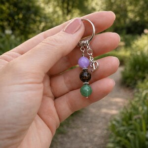 May include: A keychain with a silver ring and a charm with four gemstone beads in purple, brown, and green. A small silver charm with the letter "D" hangs below the beads. The background is a blurred outdoor scene.