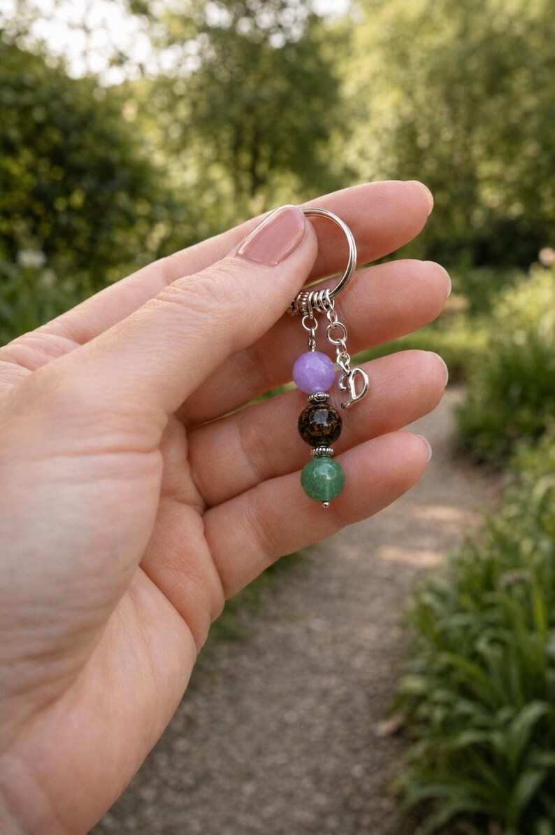 May include: A keychain with a silver ring and a charm with four gemstone beads in purple, brown, and green. A small silver charm with the letter "D" hangs below the beads. The background is a blurred outdoor scene.