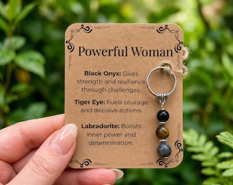 May include: A silver-toned metal ring charm featuring three spherical beads in black, brown, and grey, attached to a rectangular kraft paper card with the text Powerful Woman and descriptions of Black Onyx, Tiger Eye, and Labradorite.