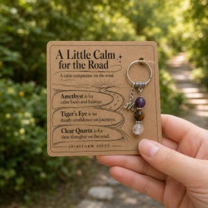 May include: A metal keyring featuring three round gemstone beads—amethyst, tiger's eye, and clear quartz—and a silver letter M charm, attached to a tan card labelled A Little Calm for the Road with descriptions of the stones and Spiritgem Gifts.
