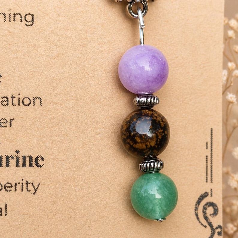May include: A beaded pendant featuring three spherical gemstones in purple, brown, and green. The beads are separated by silver-colored metal accents and suspended from a silver-colored hook. The pendant is displayed against a tan background with printed text.