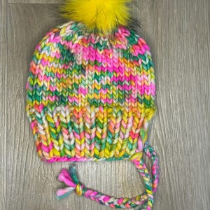 Könnte beinhalten: Eine bunte Strickmütze mit einem gelben Bommel und langen geflochtenen Bändern. Die Mütze ist aus verschiedenen Farben gefertigt, darunter Pink, Grün, Gelb und Orange.