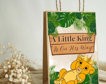 May include: A brown paper gift bag with twisted paper handles. The bag features a cartoon lion cub wearing a gold crown, surrounded by green tropical leaves. The text "A Little King Is On His Way" is displayed on a wooden sign.
