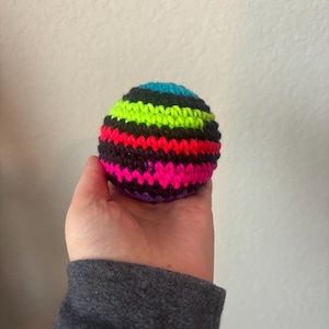 May include: A colorful, striped crochet ball in shades of black, neon green, pink, purple, blue, and teal. The ball is held in a hand against a plain white background. The ball is a sphere.