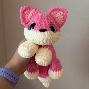 May include: A handmade pink and white crocheted fox plush toy with large, glittery eyes. The fox has a fluffy texture, with pink ears, head, and body, and white accents on the face, paws, and tail. A cute and cuddly stuffed animal.