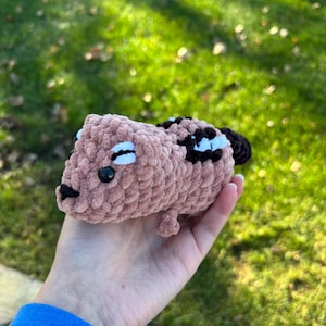 May include: A handmade, plush, crochet animal in shades of pink, brown, black, and white. The small, rounded creature has black eyes, a black nose, and white accents. The toy is held in a hand against a background of green grass.