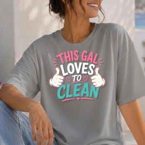 以下が含まれることがあります： ピンク、白、ターコイズブルーの文字で「THIS GAL LOVES TO CLEAN」と書かれたグラフィックプリントが施されたグレーの半袖Tシャツ。デザインには、親指を立てた漫画風の手のイラストと小さなピンクのハートのアイコンが含まれています。