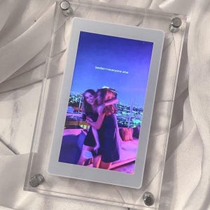 May include: A clear acrylic photo frame with a white border, displaying a digital image. The image shows two people hugging, with the text "bestie>>>everyone else". The background is a nighttime cityscape.