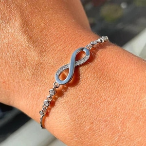 May include: A silver infinity symbol bracelet with small clear cubic zirconia stones. The bracelet is on a person's wrist.