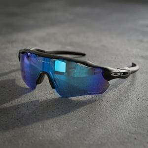 Könnte beinhalten: Schwarze Sportsonnenbrille mit blau verspiegelten Gläsern. Die Sonnenbrille hat einen gebogenen Rahmen und der Markenname "Oakley" ist oben am Rahmen sichtbar. Die Gläser sind leuchtend blau.
