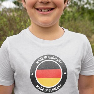 Pode incluir: Uma camiseta branca com um emblema circular com as cores da bandeira alemã: preto, vermelho e amarelo. O texto "MADE IN GERMANY" circunda o emblema. A camiseta é usada por uma pessoa.