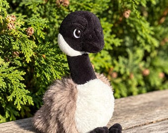 May include: A plush Canada goose toy featuring a black head and neck, a white cheek patch, a white chest, and light brown fuzzy wings. The stuffed animal has embroidered eyes and small black feet, designed with a soft, textured fabric finish.