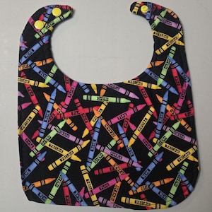 Könnte beinhalten: Ein schwarzes Baby-Lätzchen mit einem bunten Buntstiftmuster. Das Lätzchen zeigt eine Vielzahl von Buntstiftfarben, darunter Rot, Gelb, Blau, Grün und Lila, die über die Oberfläche verteilt sind. Zwei gelbe Druckknöpfe sichern das Lätzchen.
