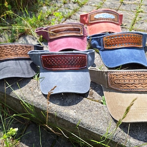 Könnte beinhalten: Eine Sammlung von fünf Sonnenblenden in verschiedenen Farben, darunter Anthrazit, Kastanienbraun, Blau, Rot und Hellbraun. Jede Blende verfügt über ein braunes Lederpanel auf der Vorderseite, das mit einzigartigen Prägemustern und Nähten verziert ist.
