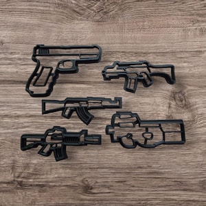 May include: Five black metal cookie cutters shaped like different types of guns. The cutters are on a wooden surface.