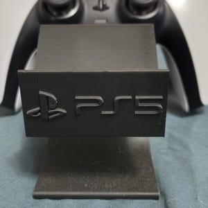 Puede incluir: Un soporte negro para consola PS5 con el logotipo PS5 en relieve en la parte delantera. El soporte está hecho de un material negro mate y está diseñado para sostener una consola de juegos. Un mando de juego blanco y negro está en el fondo.