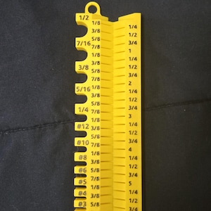 May include: A yellow, foldable drill bit gauge with measurements in inches. The gauge has a series of cutouts and markings for various drill bit sizes, including fractions like 1/8, 3/8, and 1/2, and numbered sizes.