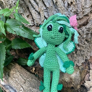 May include: A handmade green crochet fairy doll with large black eyes, light blue wings, and a pink flower accent. The fairy is posed on a textured, brown tree trunk, surrounded by green leaves. The doll is approximately 6 inches tall.