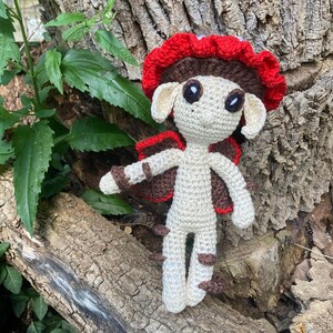 May include: A handmade crochet mushroom fairy doll. The doll has a cream-colored body, brown and red wings, and a red and brown mushroom cap. The doll is posed on a tree trunk.