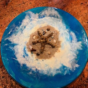 May include: A round, ocean-themed resin art piece. The design features a sandy island with a gold starfish, surrounded by blue and white resin waves. Small shells and stones are scattered on the island.