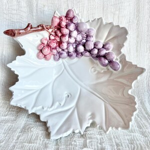 May include: A white ceramic serving dish shaped like a leaf, with a cluster of grapes in shades of pink and purple. The grapes are attached to a brown stem. The dish is on a white textured surface.