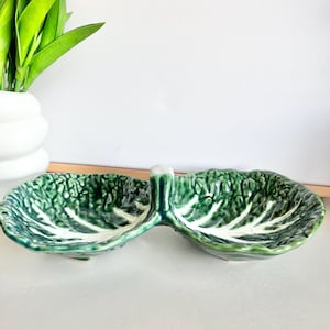 Puede incluir: Un plato de servir de cerámica verde y blanco con forma de hoja de col. El plato tiene dos secciones y un asa en el centro.