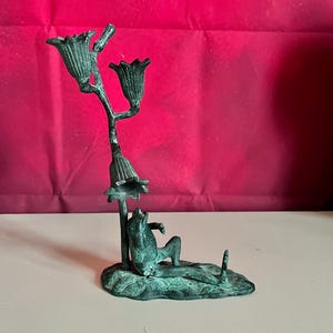 May include: A bronze-colored frog figurine lounging beneath a floral candelabra. The frog rests on a lily pad base, with the candelabra featuring three flower-shaped candle holders. The sculpture has a weathered, antique appearance.